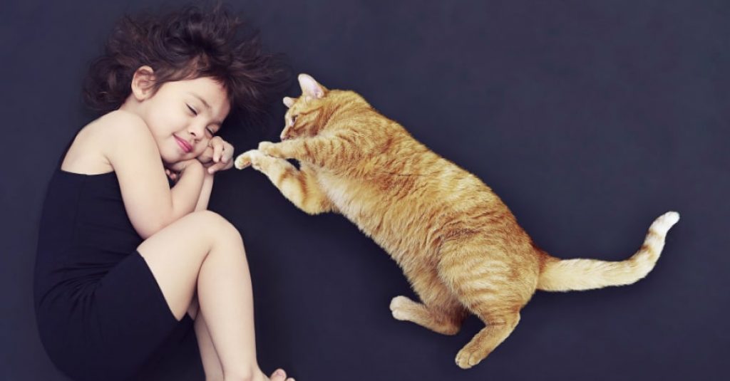 the baby girl lies on the bed with dark sheets and a ginger cat lies near her